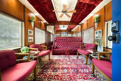 Interior of a cozy lounge with red velvet furniture, wooden walls, and a patterned rug, featuring a ceiling fan and large windows.