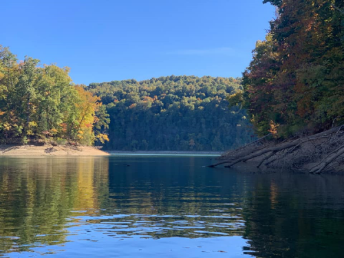 A serene lake surrounded by trees in autumn colors, reflecting the clear blue sky and distant hills.