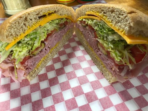 A freshly cut sandwich with layers of ham, cheese, lettuce, and tomato on whole grain bread, served on a checkered paper.