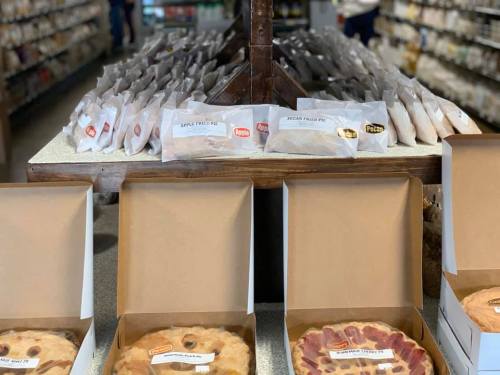 A display of various pies and packaged desserts on a table in a store, with labels for apple and pecan pies.