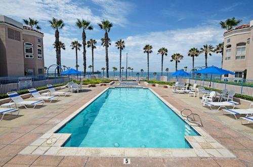 A clear swimming pool surrounded by lounge chairs and palm trees, with a beach and ocean in the background.