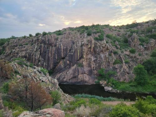 A rocky cliffside with greenery and a calm river below, under a cloudy sky at sunset.
