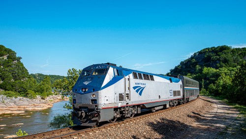 Amtrak train traveling along a riverbank, surrounded by lush greenery and blue skies.