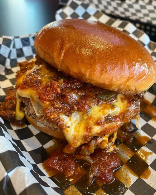 A close-up of a juicy burger topped with melted cheese and chili, served on a shiny bun with a checkered paper background.