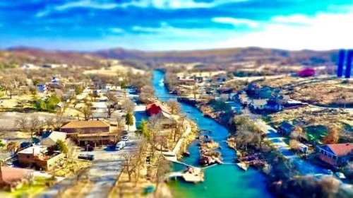 Aerial view of a scenic river winding through a small town, surrounded by trees and hills under a bright blue sky.