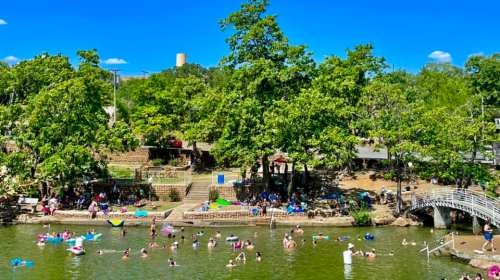 A sunny park scene with people swimming and relaxing in a river, surrounded by trees and a bridge in the background.