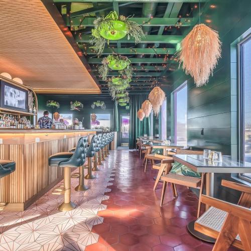 A stylish restaurant interior featuring a bar, green decor, hanging plants, and modern seating arrangements.
