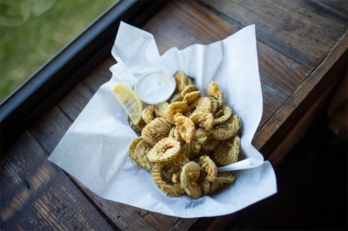 A basket of crispy fried pickles with a side of dipping sauce and a lemon wedge, resting on a wooden table.