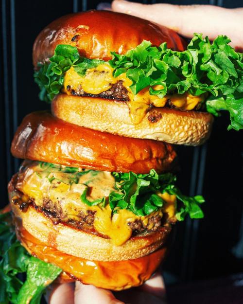 A towering double burger with melted cheese and fresh lettuce, held by a hand against a dark background.