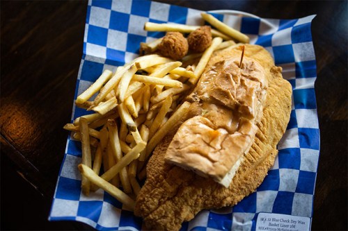 A plate with fried fish, a sandwich, hushpuppies, and a side of French fries on a blue checkered paper.