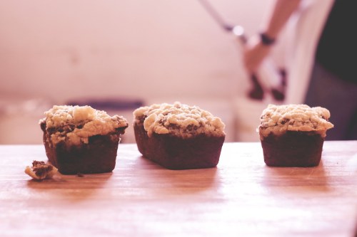 Three small loaves of baked goods with crumbly tops on a wooden surface, with a blurred figure in the background.