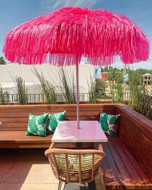 A vibrant pink umbrella shades a table on a wooden patio, surrounded by green plants and tropical-patterned cushions.