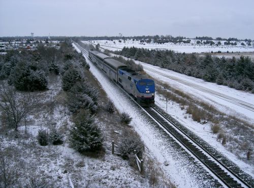 A train travels along snowy tracks, surrounded by trees and a winter landscape under a cloudy sky.