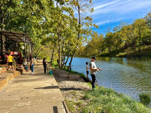 A peaceful riverside scene with people fishing and enjoying nature on a sunny day. Trees line the water's edge.