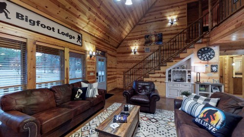 Cozy lodge interior with wooden walls, leather sofas, a coffee table, and a staircase, featuring a Bigfoot Lodge sign.
