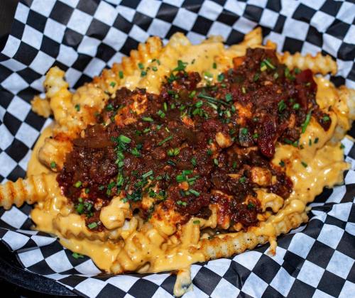 A serving of crinkle-cut fries topped with cheese sauce, chili, and chopped chives, served in a black and white checkered basket.