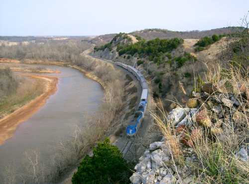 A train travels along a curved track beside a river, surrounded by hilly terrain and sparse trees.