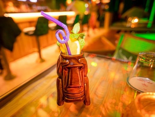 A colorful tiki mug filled with a tropical drink, garnished with mint and a lemon slice, sits on a wooden table.