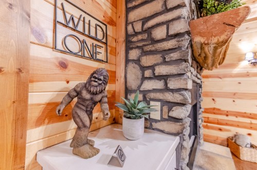 A wooden figure of Bigfoot stands next to a potted plant in a rustic room with wooden walls and a stone fireplace.