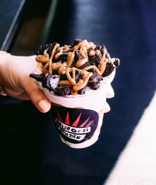 A hand holding a cup filled with dessert topped with crushed cookies and drizzled with caramel sauce.