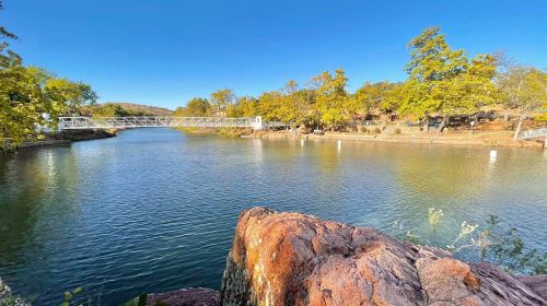 A serene river scene with a bridge, surrounded by trees and rocky banks under a clear blue sky.