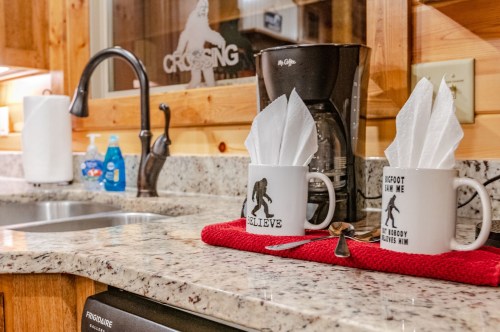 Two mugs with Bigfoot designs sit on a granite countertop, alongside a coffee maker and neatly arranged napkins.