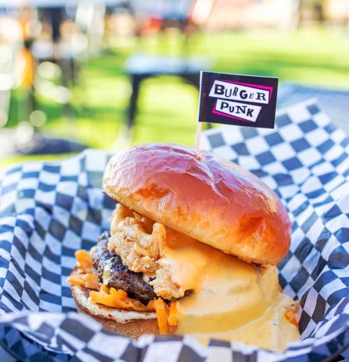A close-up of a burger with a golden bun, crispy chicken, cheese sauce, and a "Burger Punk" flag on top, served in a checkered wrapper.