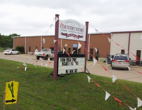 Sign for a country store advertising fresh produce, vegetables, and ice, with cars parked nearby and decorative flags.