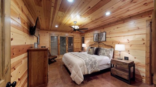 Cozy bedroom with wooden walls, a queen bed, nightstands, a TV, and a ceiling fan, featuring warm lighting.