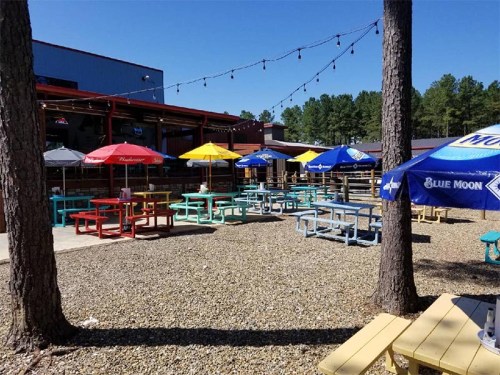 Outdoor seating area with colorful picnic tables and umbrellas, surrounded by trees and string lights.