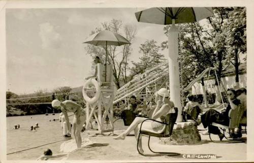A vintage black-and-white photo of a swimming area with people relaxing, a lifeguard, and a water slide.