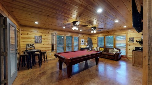 Cozy game room with a pool table, wooden walls, and large windows, featuring a sofa and bar stools.