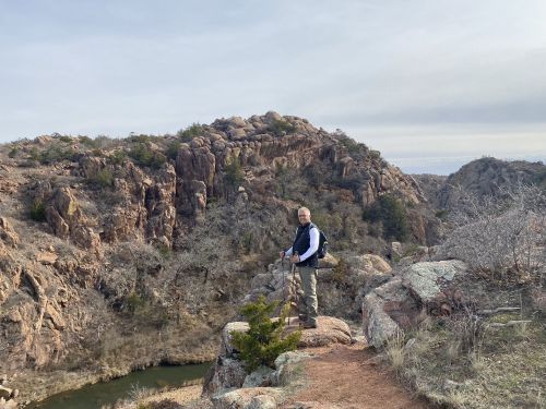 A person stands on a rocky ledge overlooking a scenic canyon with a river below and hills in the background.
