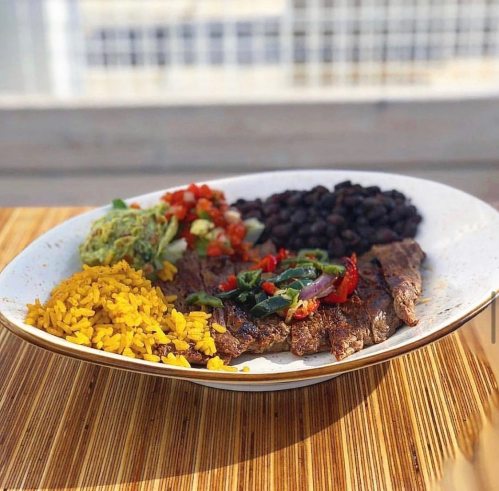 A plate of grilled steak with yellow rice, black beans, fresh salsa, and guacamole on a wooden table.