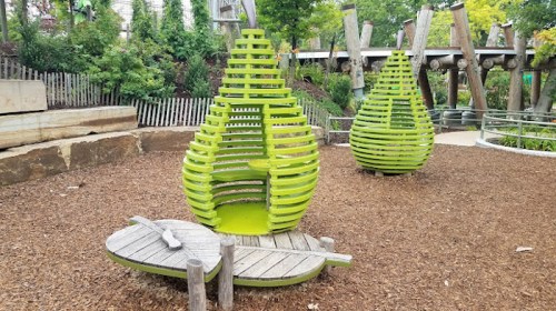 Two green, pear-shaped structures in a park, surrounded by wood chips and greenery, with a wooden boat nearby.