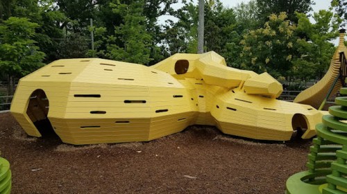 A large, abstract yellow playground structure with various openings, surrounded by green trees and wood chips.