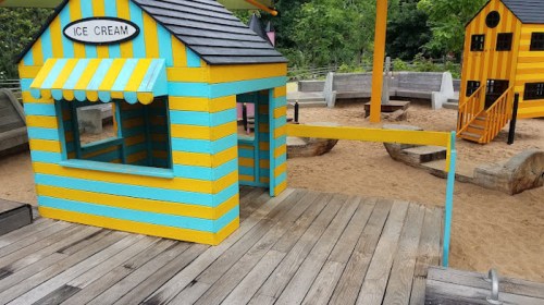 Colorful ice cream stand with blue and yellow stripes, surrounded by a sandy play area and wooden structures.
