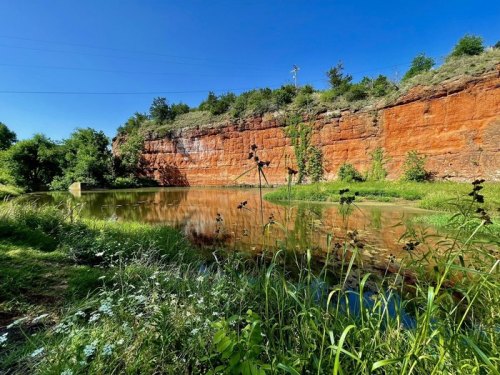 A serene pond surrounded by lush greenery and a red rock cliff under a clear blue sky.