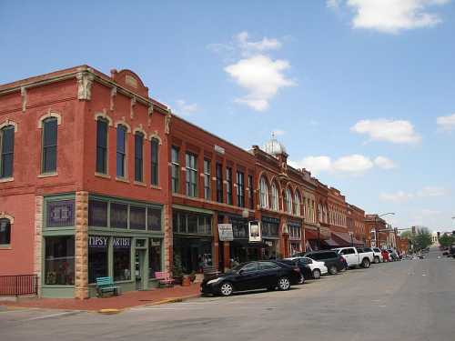 A charming street lined with red brick buildings, shops, and parked cars under a blue sky with fluffy clouds.