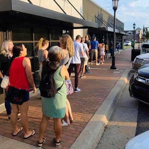 A long line of people waiting outside a storefront on a sunny day, with cars parked nearby.