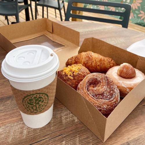 A coffee cup next to a box of assorted pastries, including a croissant, doughnut, and cinnamon roll, on a wooden table.