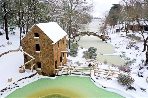 A snowy landscape featuring a stone building by a green pond and a bridge over a river, surrounded by trees.