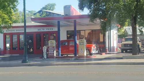 A retro diner with a red car parked outside, featuring vintage gas pumps and a sign that says "Burgers & Fries Now Open."