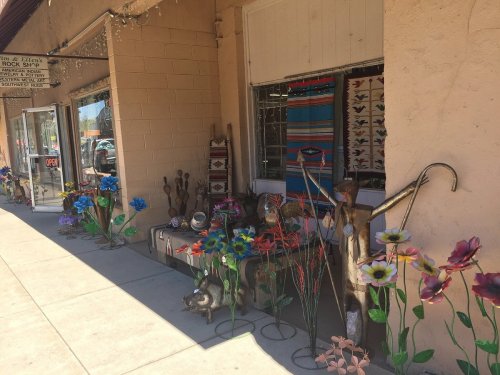 Colorful metal flowers and sculptures decorate a storefront, with a sign indicating the shop is open.