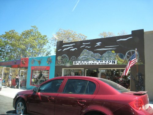 A red car parked in front of the Black Canyon Trading Post, featuring colorful storefronts and a clear blue sky.