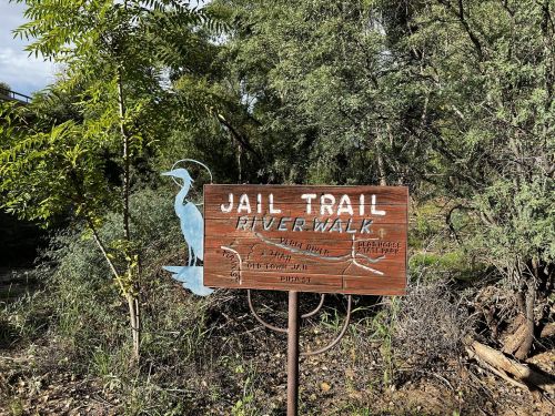 Wooden sign for "Jail Trail River Walk" with a blue bird silhouette, surrounded by greenery.