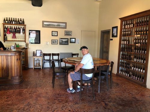 A man sits at a round table in a cozy wine tasting room, surrounded by bottles and framed photos on the walls.
