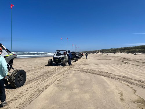 A group of off-road vehicles parked on a sandy beach under a clear blue sky, with ocean waves in the background.