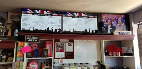 A bakery menu displayed on a screen above a counter, with a TV showing a crowd in the background.