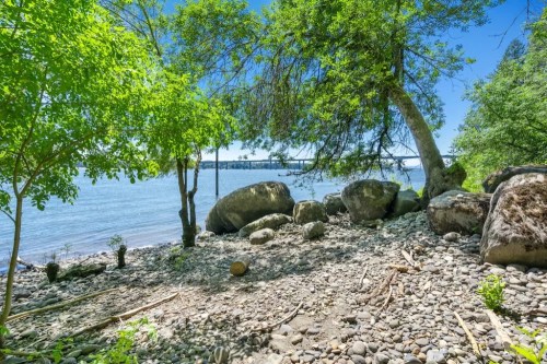 A serene riverside scene with large rocks, greenery, and a clear blue sky reflecting on the water.